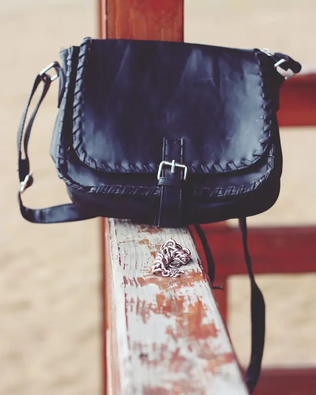 Un sac bandoulière en cuir noir à surpiqûres, posé sur une barrière en bois.